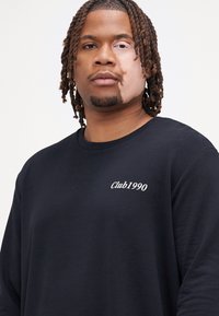 Czarny sweatshirt z haftem "Club1990" w białym kolorze na klatce piersiowej. Długie rękawy, okrągły dekolt i luźny krój. Gładka faktura.