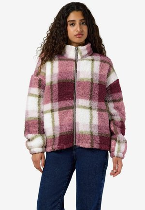 Femme portant une veste duveteuse zippée avec un motif à carreaux rose, blanc, bordeaux et vert et un jean bleu foncé, debout sur fond blanc.