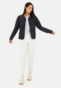 Gestepptes schwarzes Jacket mit runder Kragen und Druckknöpfen, getragen über einem weiß- und marineblau gestreiften Hemd, kombiniert mit cremefarbenen Hosen und Sneakers.