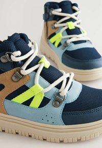 Hög-top sneakers med en blandning av marinblått, ljusblått och beige material. Texturerad mesh-överdel, kontrasterande gula accenter och vita snören.