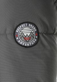 Gray fabric featuring a round embroidered patch with "Respect Brand Adventure," a mountain design, and the year "1997" in red accents.