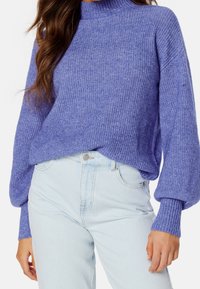 Pull violet en maille avec un col montant, une texture côtelée et des manches larges, associé à un jean bleu clair taille haute.