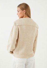 Beige stickad tröja med stor krage, ribbad textur, sänkta axlar och ett figursytt fåll. Mjuk tygkvalitet och avslappnad passform.