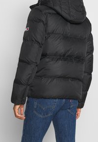 Zwarte pufferjack met capuchon, voorzien van gestikte secties, elastische manchetten en een klein logo-opdruk op de linkerschouder.