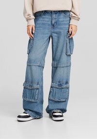 Vaaleansiniset denim-farkut, joissa on löysä istuvuus, useita etutaskuja ja ommeltu kuvio. Yhdistetty mustiin ja valkoisiin lenkkareihin.