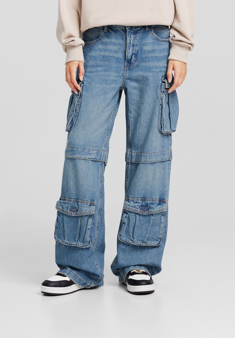 Vaaleansiniset denim-farkut, joissa on löysä istuvuus, useita etutaskuja ja ommeltu kuvio. Yhdistetty mustiin ja valkoisiin lenkkareihin.