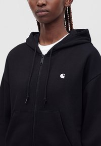Person trägt einen schwarzen Kapuzenpullover mit Reißverschluss, einem kleinen weißen Logo auf der Brust und einem weißen Shirt darunter vor einfarbigem Hintergrund.