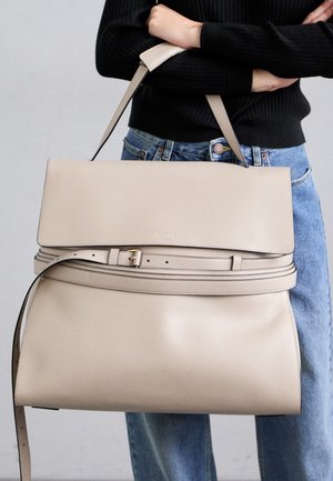 Person wearing black sweater and blue jeans holding large beige Moschino leather handbag with shoulder strap and belt detail.
