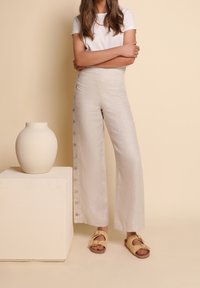 Kvinna klädd i högmidjade beige vida byxor med sidoknappar, vit kortärmad topp och beige sandaler, stående bredvid en krämfärgad keramisk vas.