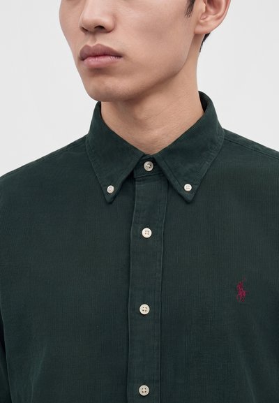 Homme portant une chemise en velours côtelé vert foncé avec boutons, col boutonné et un petit logo rouge brodé sur la poitrine.