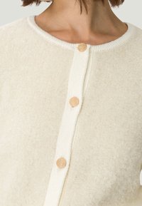 Cremfarbener Cardigan mit strukturiertem Stoff, rundem Ausschnitt und drei dekorativen Knöpfen aus einem materialähnlichen, hellen Holz.