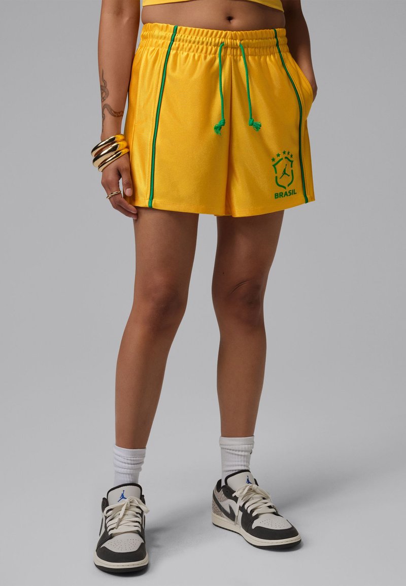 Άτομο που φοράει κίτρινα αθλητικά σορτς της Βραζιλίας με πράσινη επένδυση, ασπρόμαυρα αθλητικά παπούτσια, λευκές κάλτσες, χρυσά βραχιόλια και χέρι με τατουάζ.