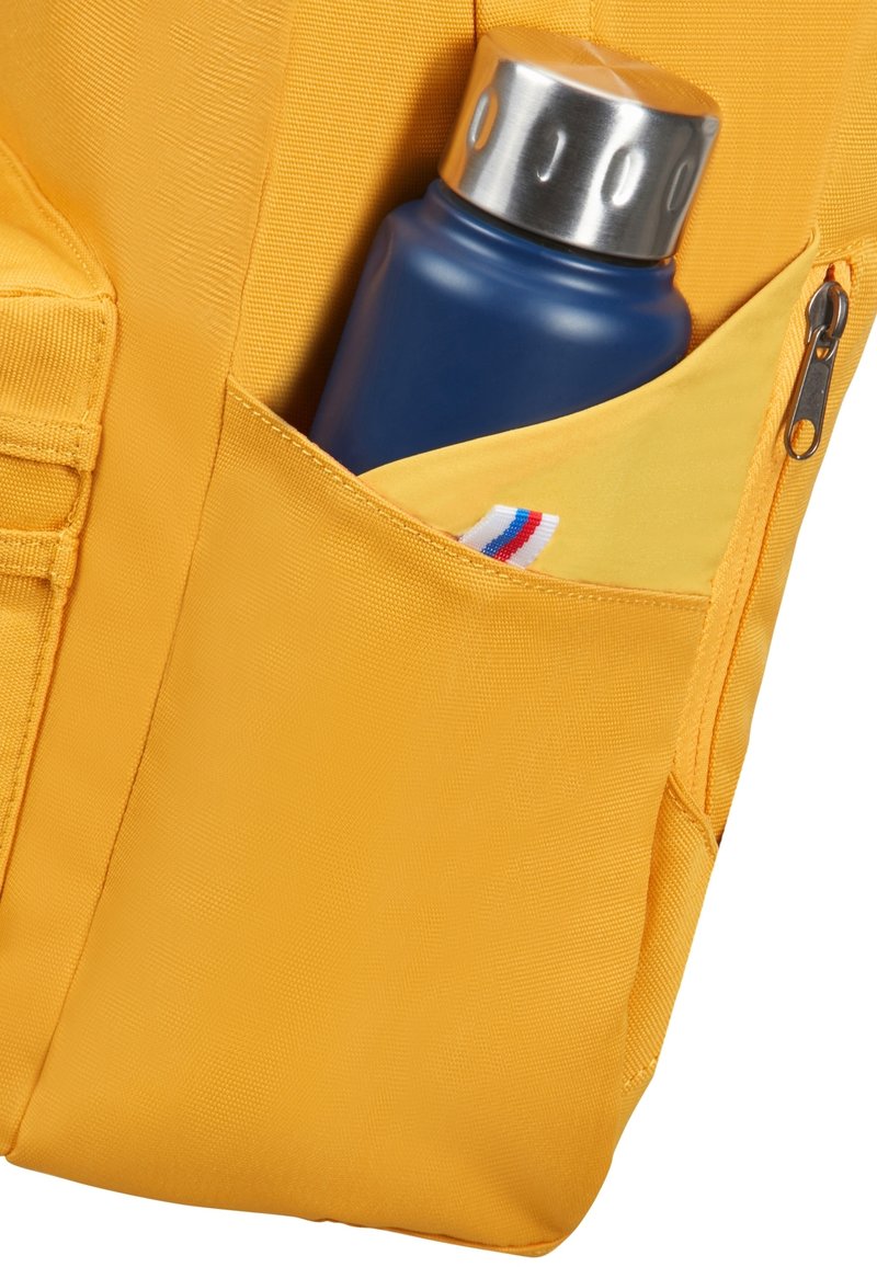 Sac à dos en tissu jaune avec une poche latérale contenant une bouteille d'eau bleue et une petite étiquette avec des rayures rouge, blanche et bleue.