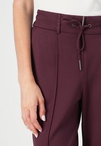 Pantalons bourgogne avec taille élastique et fermeture à cordon, présentant une texture lisse et des détails sur les coutures latérales. Positionnés à la main près de la taille.