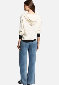 Felpa leggera crema con cappuccio e polsini neri, abbinata a jeans in denim blu a gamba larga con tasche posteriori e finitura scolorita.