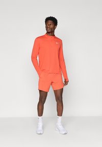 Άντρας σε πορτοκαλί αθλητικό μπλουζάκι με μακριά μανίκια και σορτσάκι, φορώντας λευκά παπούτσια και κάλτσες, στέκεται με τα χέρια στις τσέπες μπροστά σε απλή φόντο.