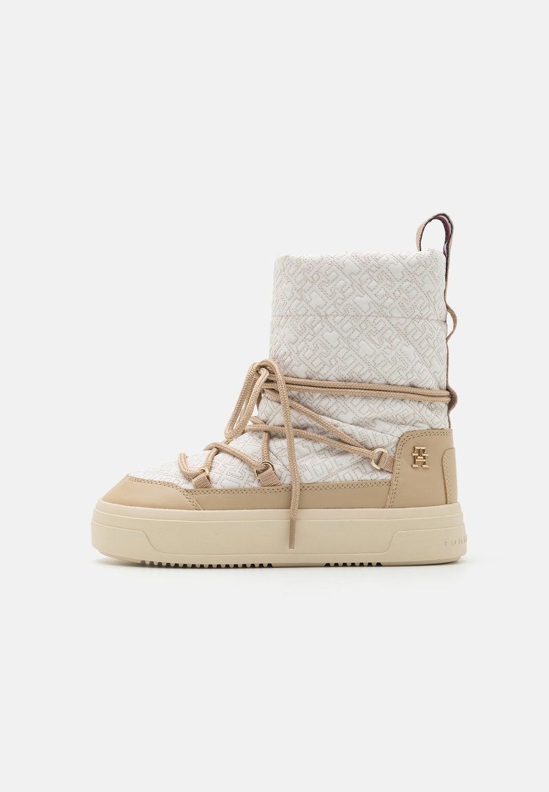 Botte d'hiver beige et blanche à tige haute avec tissu à motif, lacets enveloppants, semelle épaisse et logo "TH" doré sur le talon.