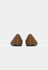 Mocassins en daim marron avec une texture lisse, une forme pointue et des semelles en caoutchouc noir. Présentent des coutures minimales et un design épuré.