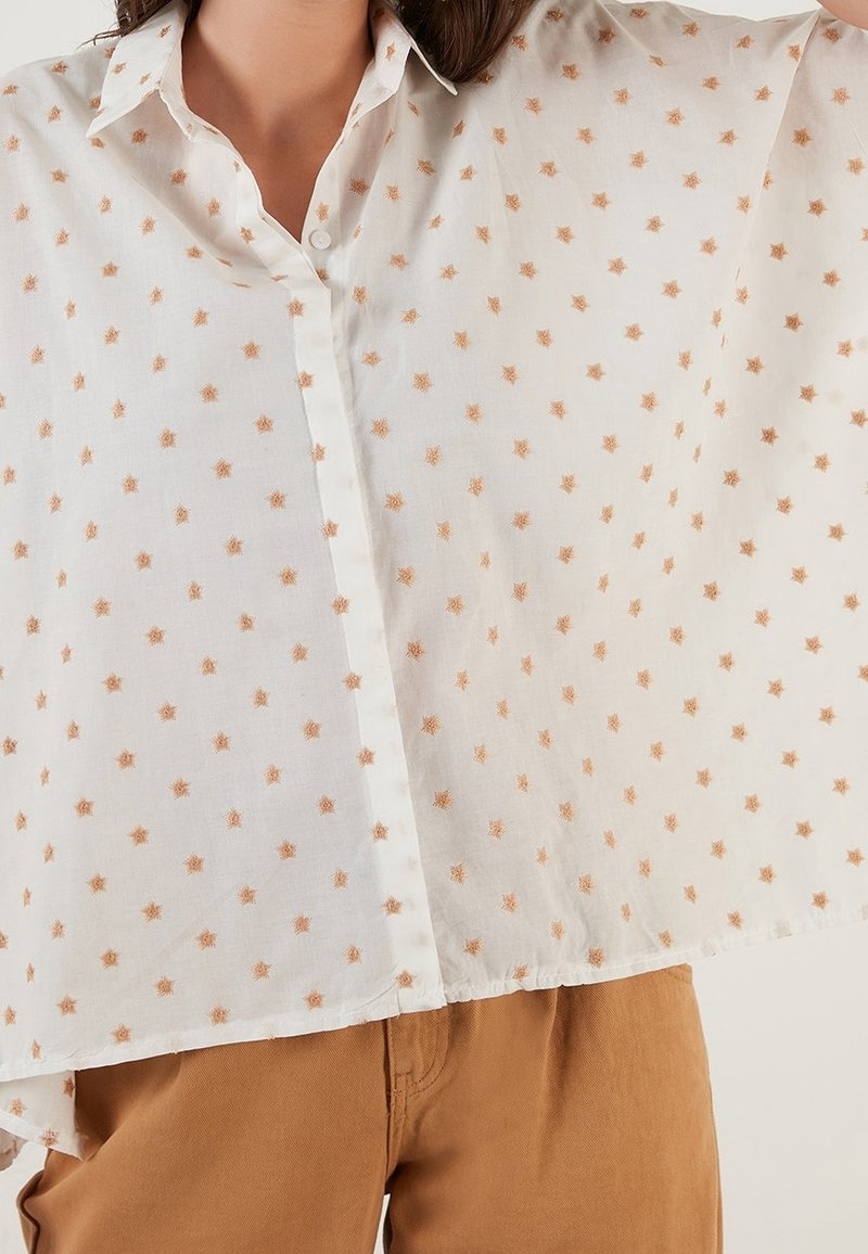 Personne portant une chemise blanche ample à boutons avec de petits motifs d'étoiles beige et un pantalon marron clair, vue de la poitrine à la hanche.