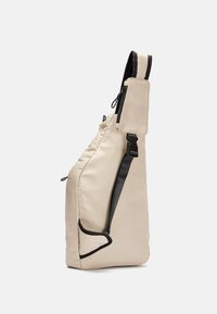 Beige nylon skulderveske med en teksturert overflate, som har et justerbart svart belte, ekstern glidelåslomme og en triangel silhuett.