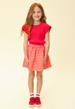 Jong meisje met lang rood haar dat glimlacht, draagt een rood topje met ruches aan de mouwen, een roze geruite rok, witte sokken en rode sneakers, staand.