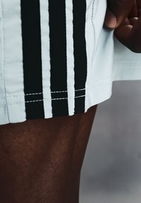 Personne à la peau foncée tenant l'ourlet d'un short blanc avec trois bandes noires verticales sur un fond gris flou.