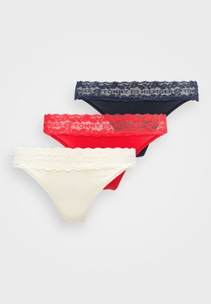 Donna con capelli ricci che indossa un reggiseno bianco e mutandine rosse con pizzo, in piedi contro uno sfondo chiaro e uniforme vicino a una sedia testurizzata.