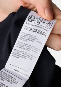 Zwarte stoffen label met verzorgingsinstructies en materiaaldetails, met tekst in meerdere talen, bevestigd aan een kledingstuk.