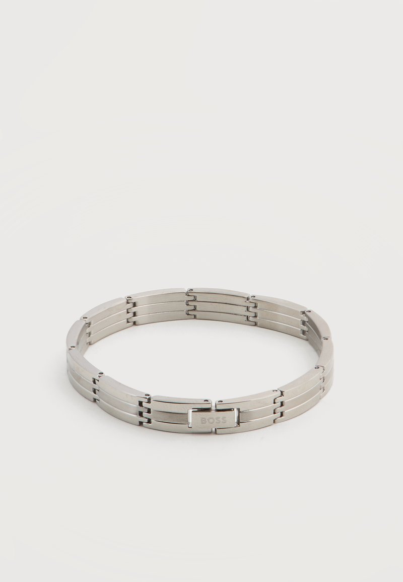 Ασημένιο μεταλλικό βραχιολάκι με εγχάρακτη αγκράφα "BOSS", τοποθετημένο σε κυκλικό σχήμα πάνω σε ένα απλό ανοιχτό φόντο.