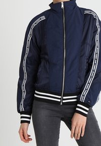 Granatowa kurtka bomberka z prążkowanymi czarnymi i białymi mankietami. Posiada zamek błyskawiczny oraz pionowe białe paski z logo wzdłuż rękawów.