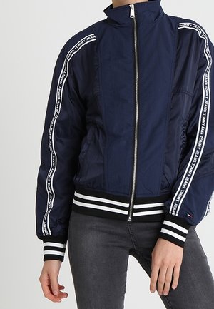 Tummansininen pomppujakku, jossa on ribbipintaiset mustat ja valkoiset hihansuut. Varustettu vetoketjukiinnityksellä ja pystysuorilla valkoisilla logoraitoja hihoissa.