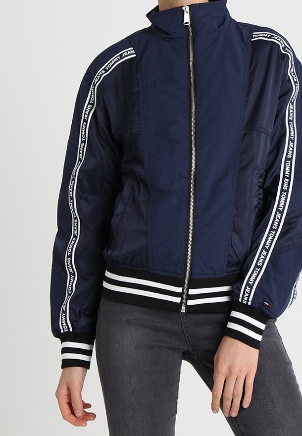 Granatowa kurtka bomberka z prążkowanymi czarnymi i białymi mankietami. Posiada zamek błyskawiczny oraz pionowe białe paski z logo wzdłuż rękawów.
