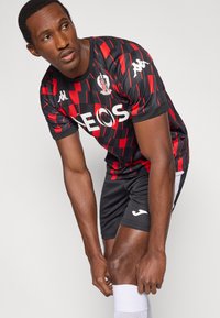 Čierno-červený športový dres s geometrickým vzorom, obsahujúci biele logo "NEOS" a značku Kappa, doplnený čiernymi šortkami a bielymi ponožkami.