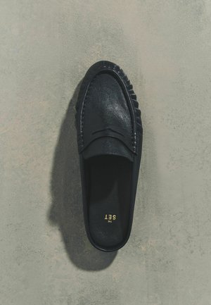 Mustast nahast paeltega jalats, millel on õmmeldud detailid ja sees kuldselt trükitud tekst "THE SET", asetatud hallile tekstuurse pinnaga alusele.