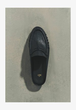 Czarny skórzany mokasyn wsuwany z dekoracyjnym przeszyciem i nadrukiem "THE SET" w złotym kolorze wewnątrz, umieszczony na szarej teksturowanej powierzchni.