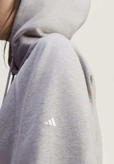 Persona que lleva una sudadera con capucha gris claro con un logo circular de Adidas by Stella McCartney en la parte superior del brazo sobre un fondo liso.
