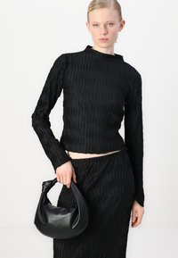 Juoda tekstūruota ilgomis rankovėmis palaidinė su aukštu kaklu, derinama su juoda plisėta sijonu. Modelis laiko mažą juodą odinę rankinę.