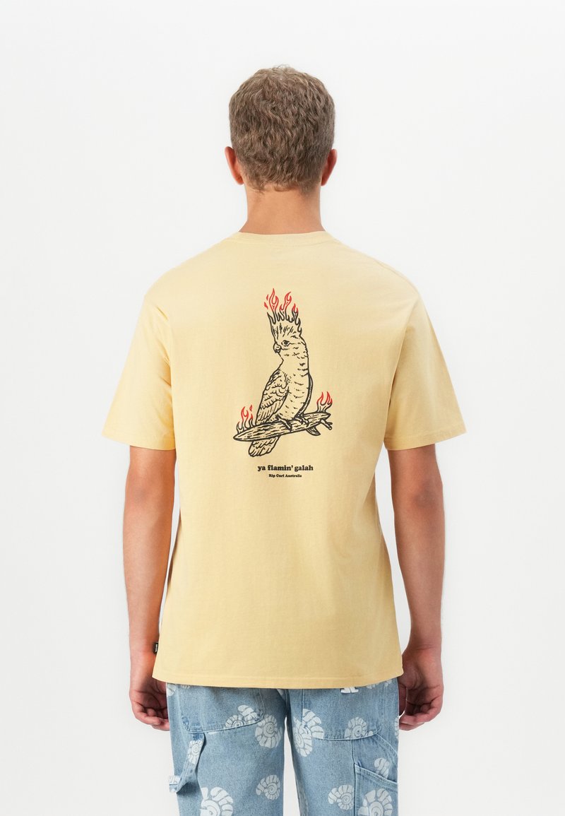 Person med gul t-shirt med grafisk sort og rød flammende kakadue på ryggen, der holder et brændende surfbræt, parret med blå mønstrede bukser.