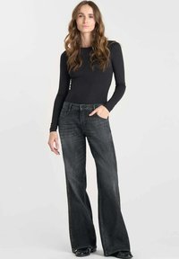 Vrouw met lang bruin haar, gekleed in een zwarte fitted long-sleeve top en een wijde, vervaagde zwarte jeans, staande met één hand in haar zak.