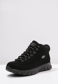 Černé vysoké tenisky s polstrovaným límcem kolem kotníku, silnou podrážkou, šněrovací konstrukcí a logem Skechers na boku, zobrazené na bílém pozadí.