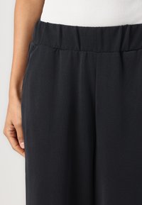 Mustad, kerged püksid elastse vöökohtaga ja sileda tekstuuriga, millel on esipoolsed voldid lisaks detailina.