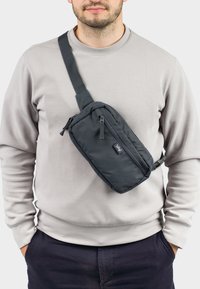 Black nylon sling bag worn crossbody, featuring a zippered pocket, adjustable strap, and rectangular shape. Smooth texture, minimal branding.