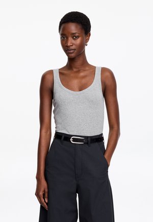 Femme aux cheveux courts portant un débardeur gris, un pantalon noir à jambes larges et une ceinture noire, debout avec une main dans la poche.
