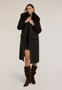 Abrigo de lana verde oscuro con cuello de piel sintética, cierre de cremallera y dos bolsillos frontales, combinado con un vestido de encaje negro y botas altas de ante marrón.