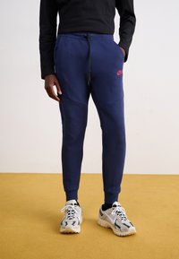 Marineblå sweatpants med elastisk talje og snoretræk, med sidelommer og et rødt logo. Satt sammen med lyse sneakers.