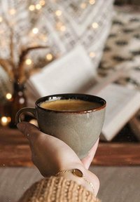 Tazza di ceramica grigia con bordo marrone, riempita di caffè. Mano che tiene la tazza, maglione lavorato a maglia caldo visibile, sfondo sfocato con luci e libro.