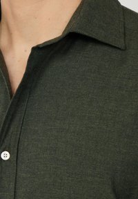 Camisa verde oscura con un tejido texturizado. Presenta un cuello puntiagudo y un solo botón blanco cerca del cuello.