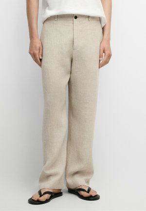 Pantaloni di lino beige indossati con sandali infradito neri, mostrati dalla vita ai piedi su uno sfondo chiaro uniforme.
