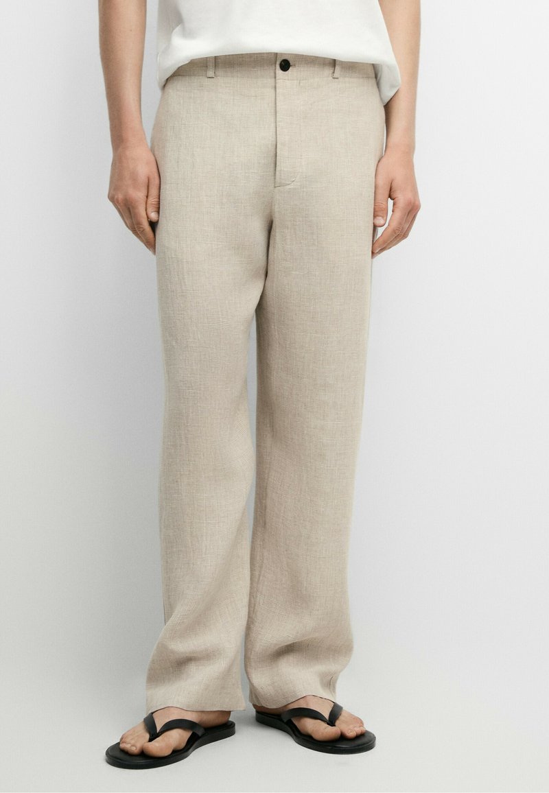 Beige linnebyxor använda med svarta flip-flops, visade från midjan till fötterna mot en enfärgad ljus bakgrund.
