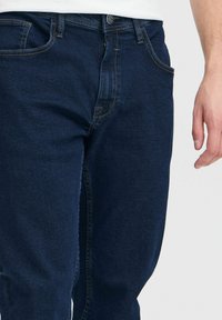 Mørkeblå denimjeans med klassisk passform, som har frontlommer, glidelås foran og kontrasterende sømmer langs sømmene.